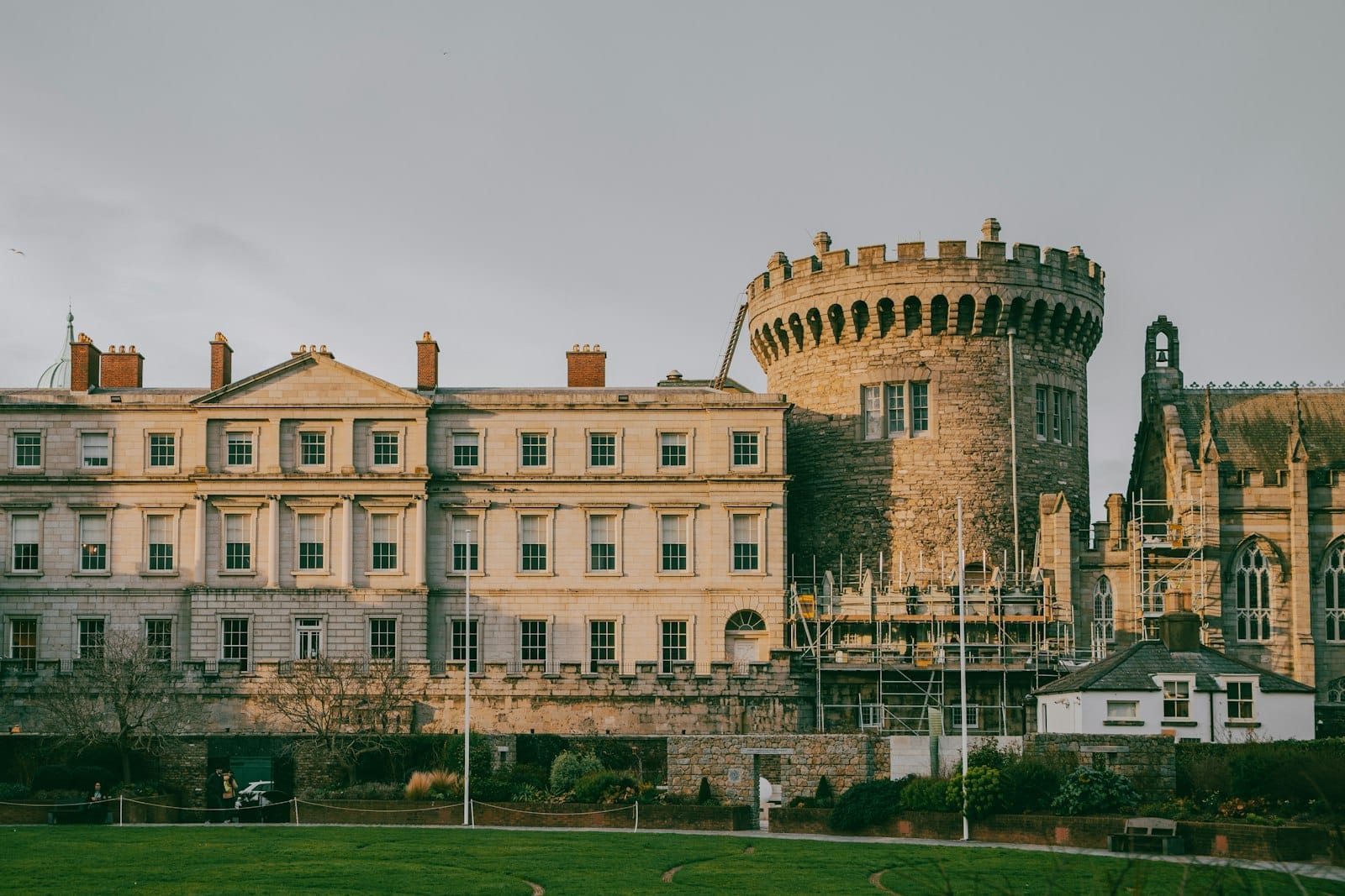 Veduta panoramica del Castello di Dublino con la torre rotonda medievale, gli Appartamenti di Stato e il prato verde circostante sotto un cielo nuvoloso.
