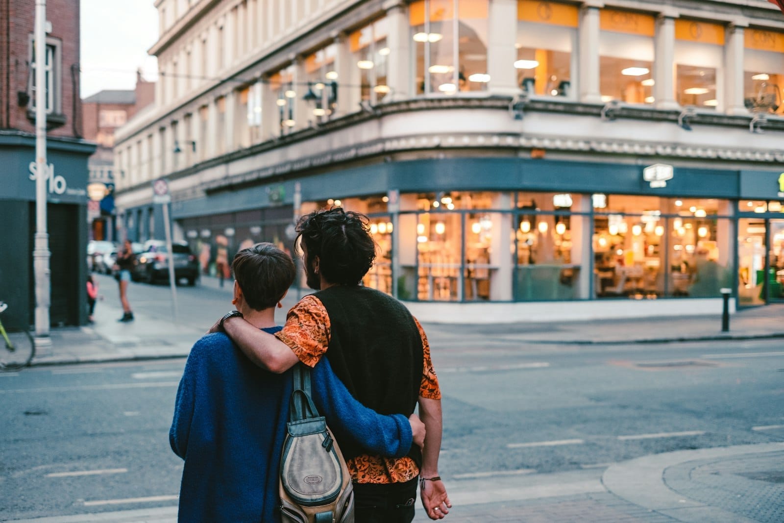 Um casal se abraça enquanto passeia por uma rua vibrante de Dublin ao entardecer, com luzes quentes nas vitrines das lojas e uma atmosfera urbana romântica.