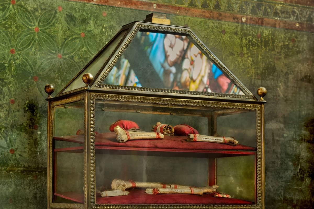 Glasreliquiar mit in rotes Tuch gehüllten Reliquien, mit einem Buntglasreflex dahinter, im Inneren einer Kirche.