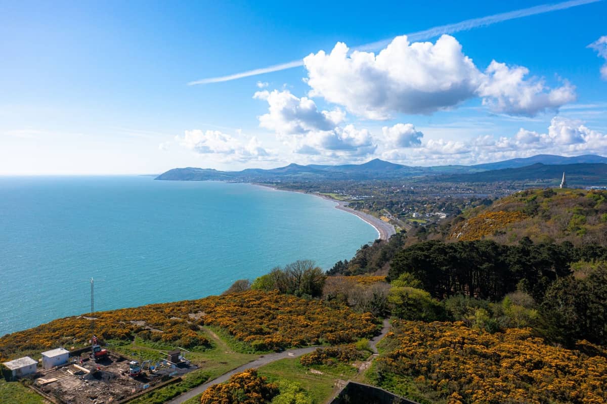 Ein weiter Küstenblick in der Nähe von Dublin mit einer dramatischen Küstenlinie, blauem Meer und üppigen Hügeln unter einem strahlend blauen Himmel mit vereinzelten Wolken.