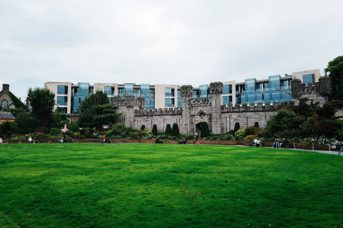 Weitblick auf den Dubh Linn Garden hinter dem Dublin Castle mit Menschen, die sich auf dem grünen Rasen entspannen, und den Burgmauern im Hintergrund.
