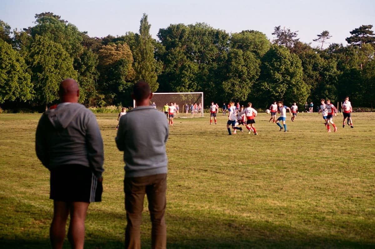 Two spectators watch a Gaelic football or hurling match played on a large grassy field with trees in the background, evoking Dublin park life.