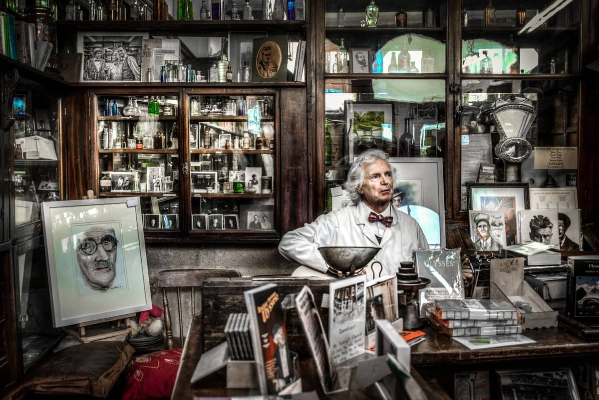 Intérieur d'une ancienne pharmacie ou boutique à thème littéraire à Dublin, avec des étagères de bouteilles et de livres, des dessins d'écrivains célèbres et une personne en blouse blanche.