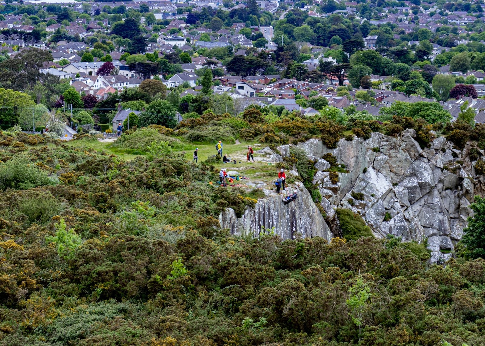 Vista de um afloramento rochoso com pessoas caminhando e escalando, cercado de vegetação, com bairros suburbanos de Dublin ao fundo.