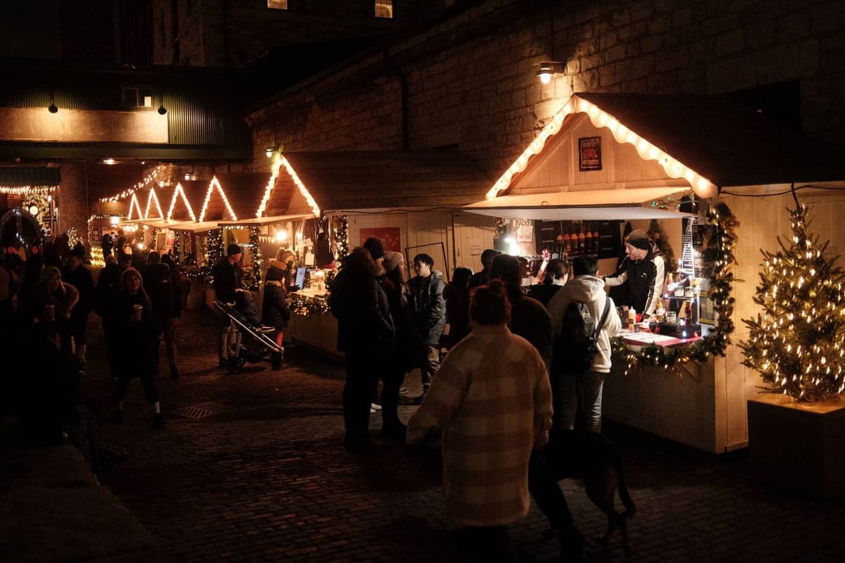 Des foules parcourant des stands en bois éclairés par des lumières festives dans un marché de Noël en plein air le soir, avec des décorations de fêtes et une atmosphère hivernale chaleureuse.