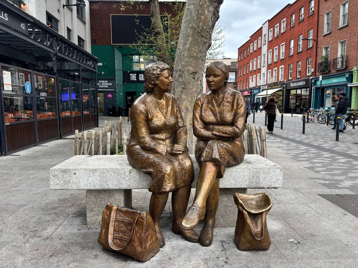 Bronzestatue zweier Frauen, die auf einer Bank in einer belebten Straße in Dublin mit Geschäften und Backsteingebäuden sitzen.