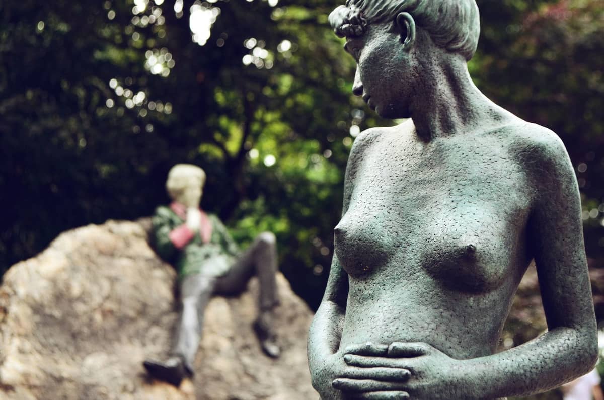 Im Vordergrund eine Bronzestatue einer Frau, im Hintergrund die farbenfrohe liegende Oscar-Wilde-Statue auf einem Felsen in einem Park in Dublin.