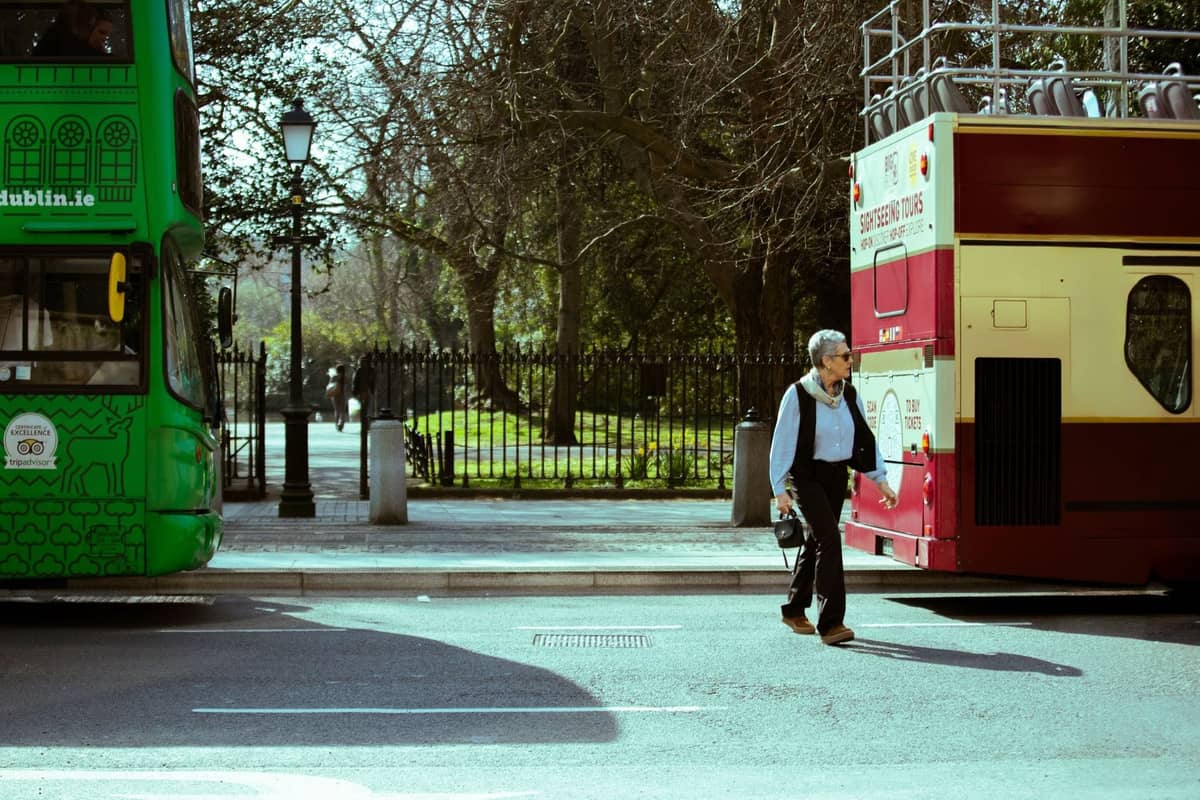 Mann, der zwischen zwei Sightseeing-Bussen auf einer Straße im Zentrum von Dublin läuft, mit Bäumen und einem Parkzaun im Hintergrund.