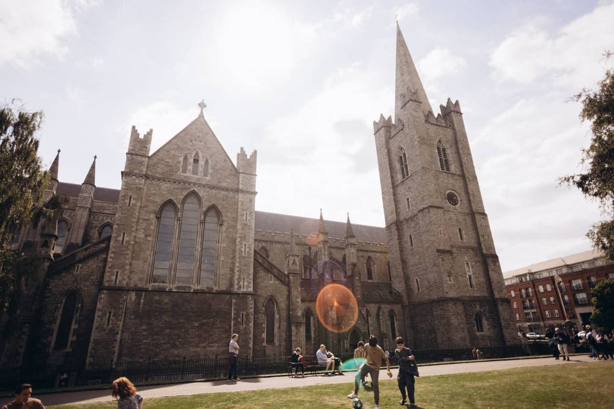 Weitwinkelansicht der St Patrick's Cathedral in Dublin mit Menschen, die tagsüber im hellen Sonnenlicht auf dem Rasen davor entspannen.
