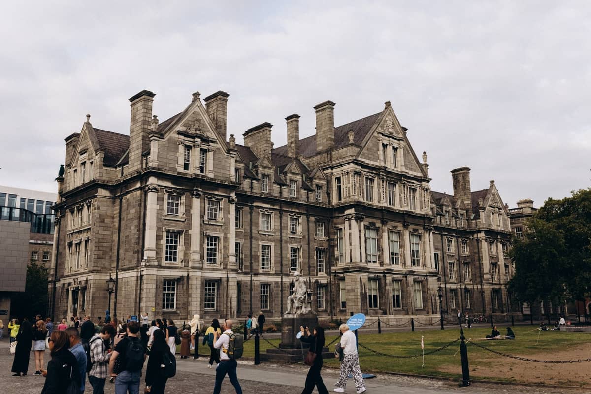 Gruppen von Menschen, die an einem bewölkten Tag vor einem prächtigen historischen Gebäude am Trinity College Dublin spazieren.