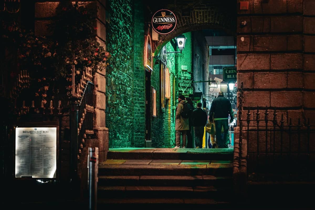 Nächtlicher Blick auf den Eingang eines Pub-Viertels in Dublin mit einem markanten Guinness-Schild und Menschen unter grünen Lichtern