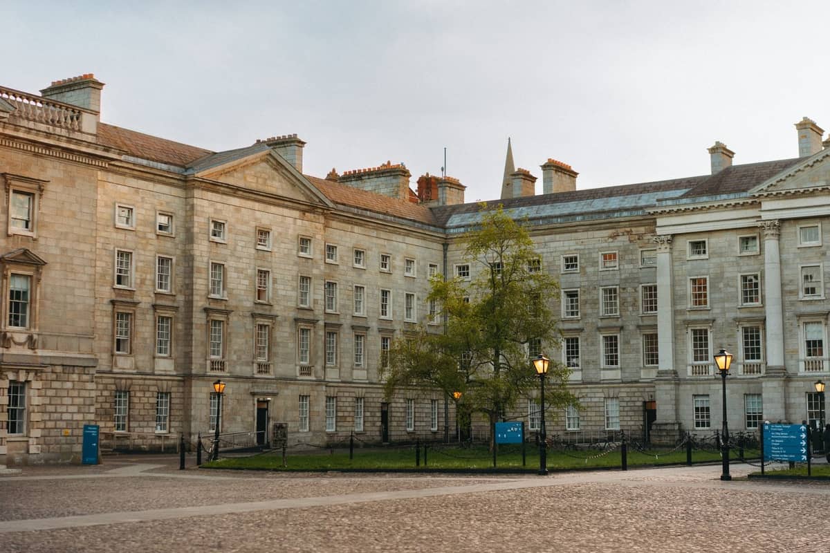 Weitwinkelaufnahme des Innenhofs des Trinity College Dublin mit historischen Steingebäuden, einem grünen Baum und blauen Informationsschildern an einem hellen Tag.