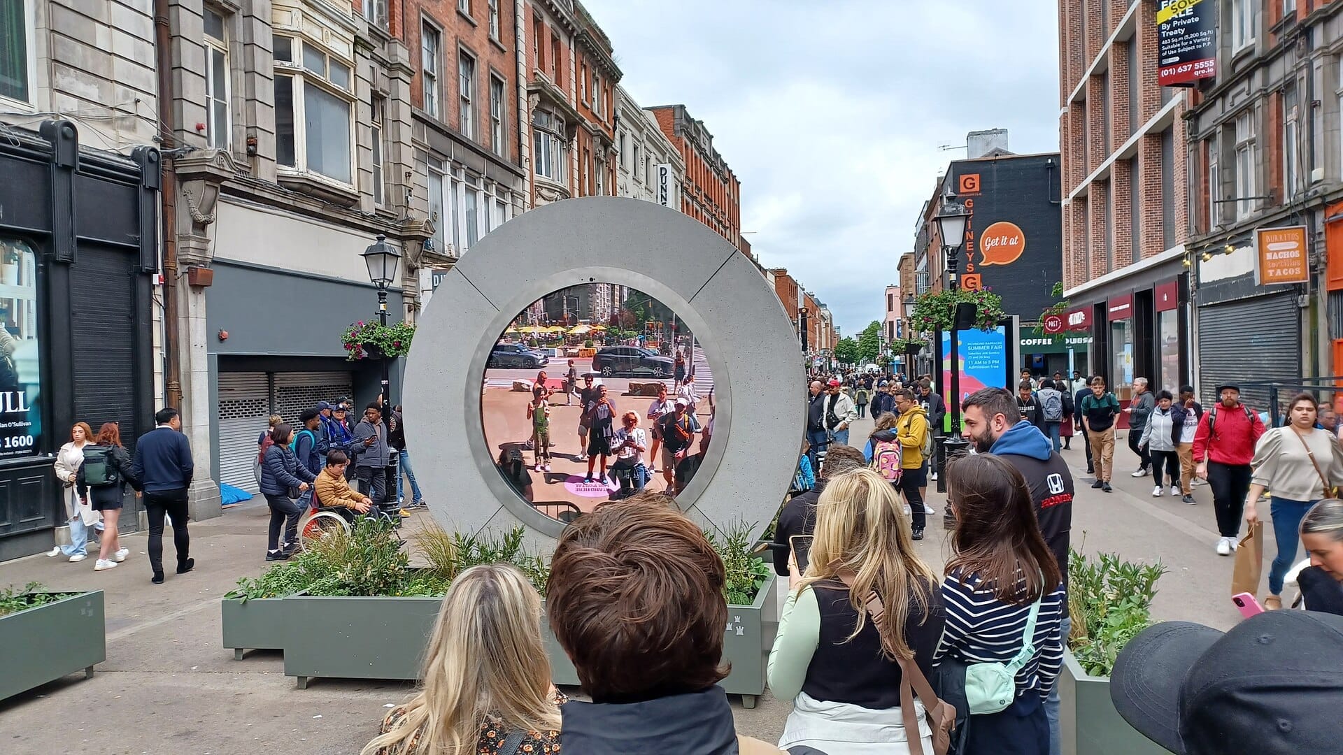Le folle si radunano su O'Connell Street per vedere la scultura circolare Dublin Portal che trasmette in diretta la vita cittadina dall'estero in una giornata trafficata e nuvolosa.