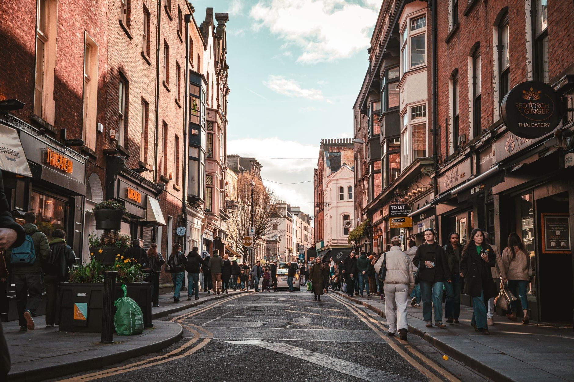 Movimentada rua de compras em Dublin com prédios de tijolos vermelhos, lojas e cafés, cheia de pessoas caminhando sob um céu parcialmente nublado.