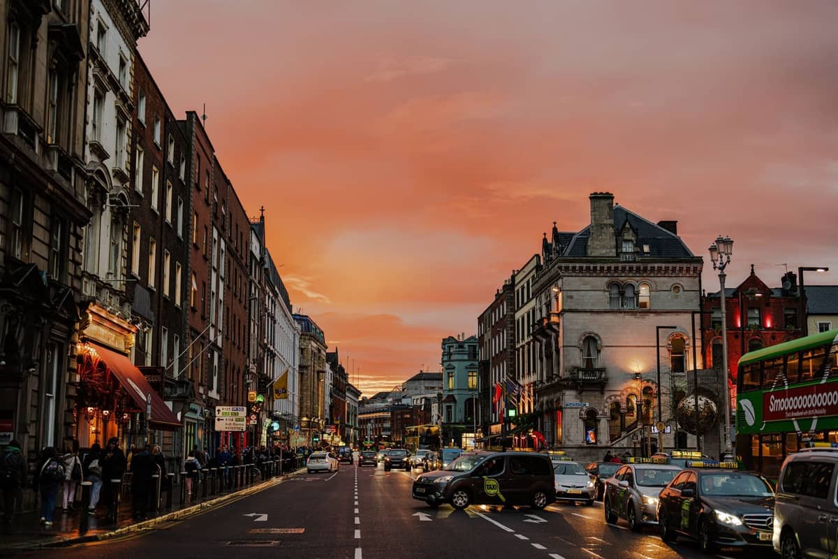 Busy Dublin street scene at sunset with shops, neon signs, pedestrians, buses and cars, showcasing the city's northside shopping area atmosphere.