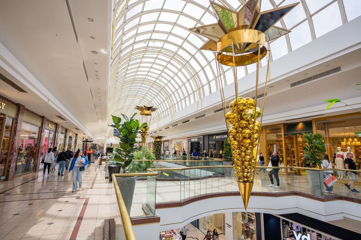 Interior view of a modern shopping mall with glass roof, shoppers walking, and stores on two levels.