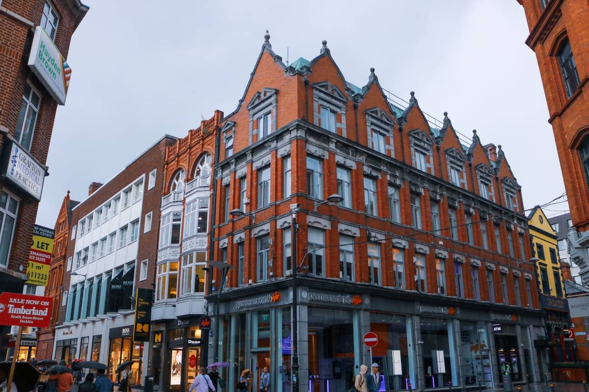 Large, elegant red-brick building on a busy pedestrian street in Dublin with shop signs and people walking by.