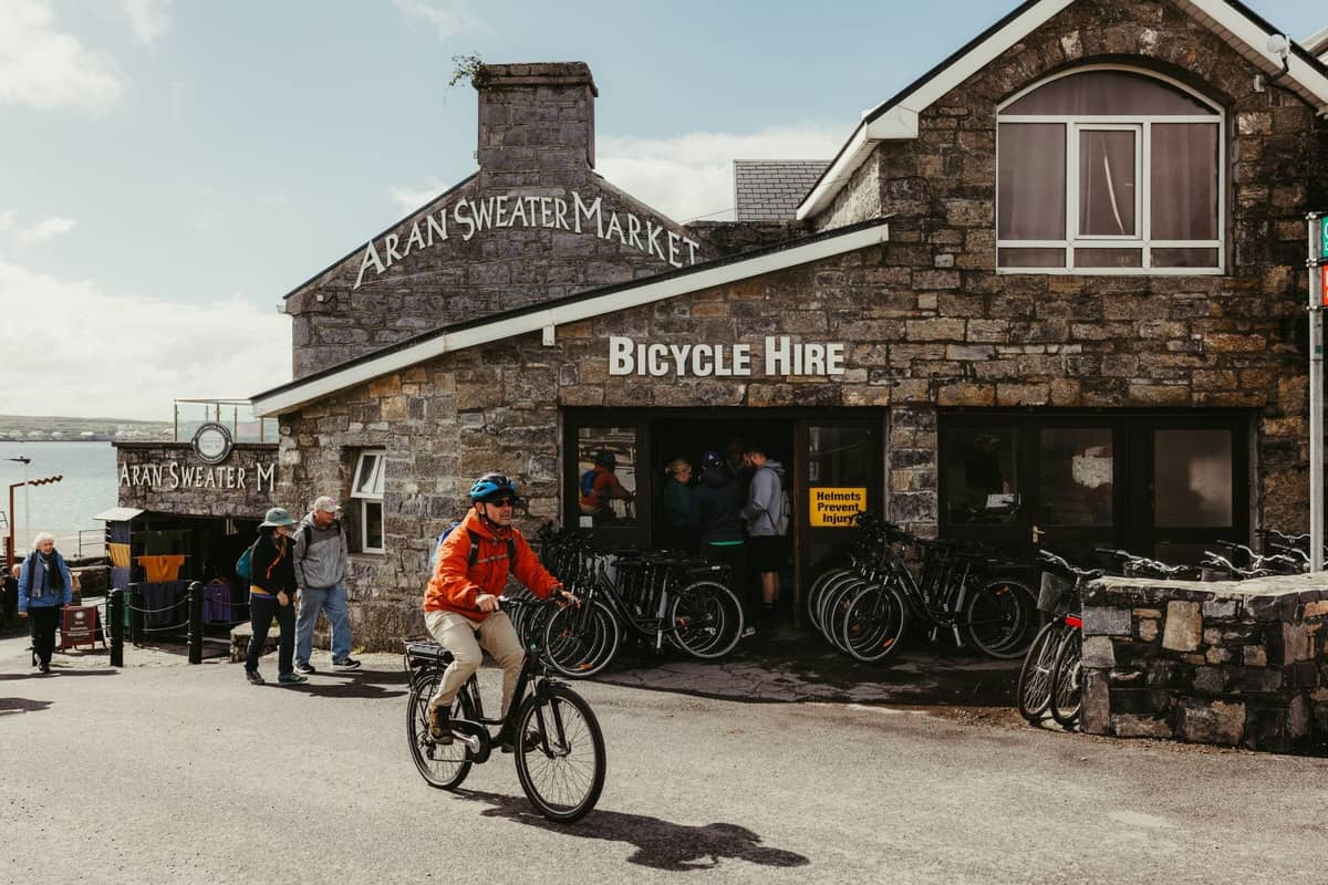 Stone building with sign 'Aran Sweater Market' in Dublin, people walking and cycling by, suggesting authentic Irish-made goods.