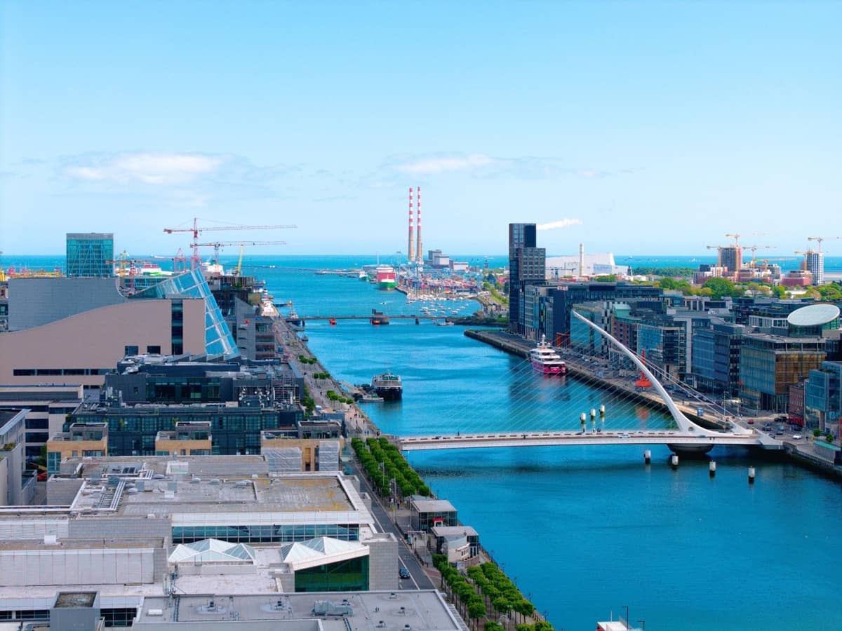 Vista aérea de los Dublin Docklands con el Samuel Beckett Bridge, edificios modernos y el río Liffey bajo un cielo azul brillante.