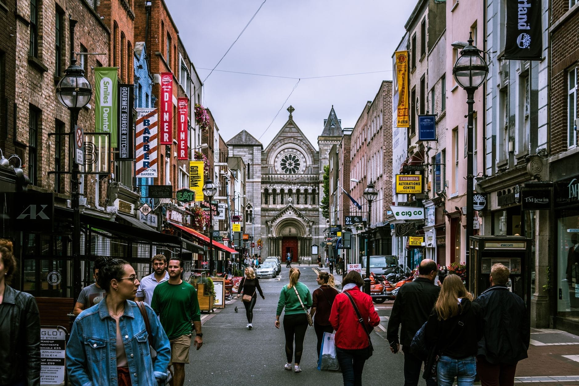 Rua movimentada de Dublin com pessoas caminhando entre lojas e pubs, letreiros coloridos e uma igreja histórica ao fundo.