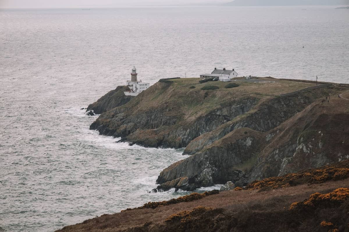 Promontorio costero con un faro y una cabaña con vistas al mar, acantilados dramáticos y olas, típico de los paseos costeros de Howth en Dublín.