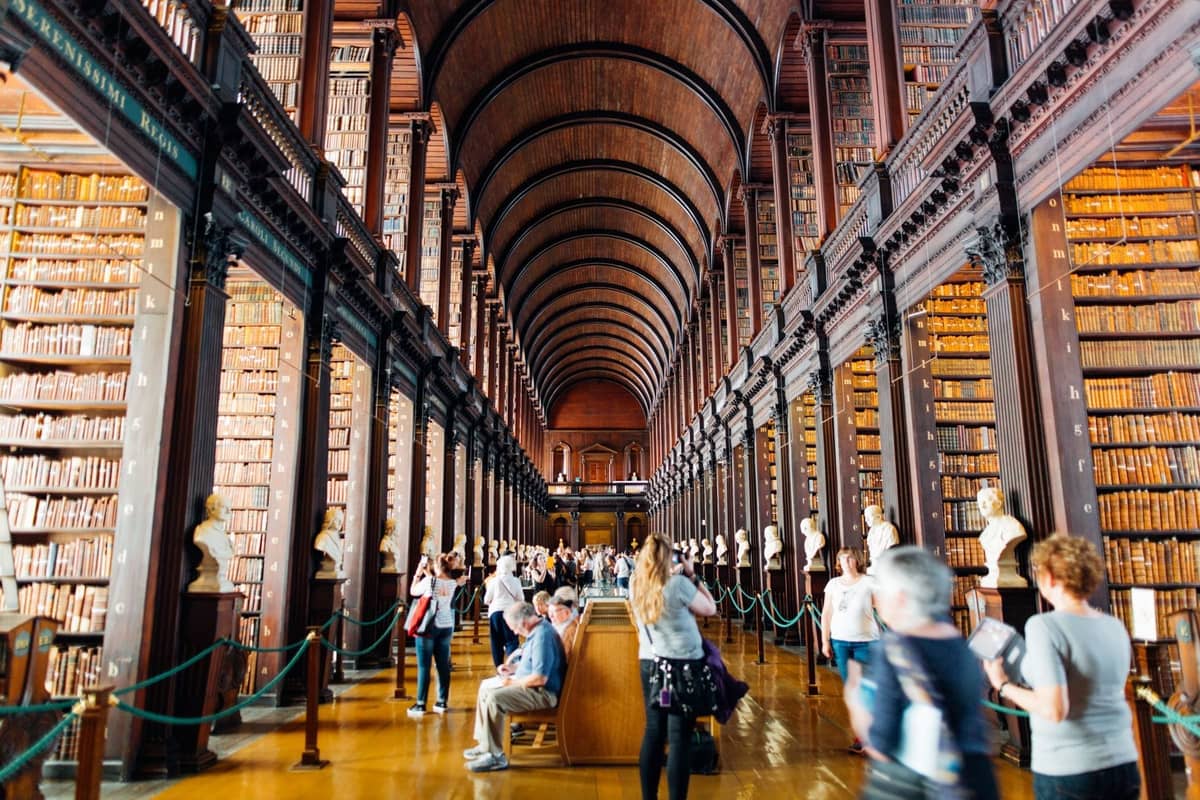 La Long Room de la Biblioteca del Trinity College en Dublín, llena de visitantes, altas estanterías y bustos clásicos a lo largo del pasillo.