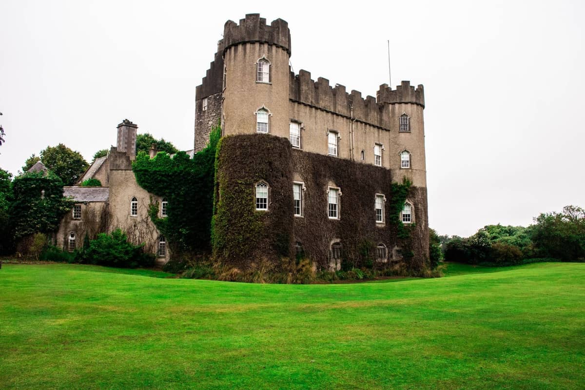 Un histórico castillo cubierto de hiedra con torres almenadas rodeado de amplios jardines verdes bajo un cielo nublado.