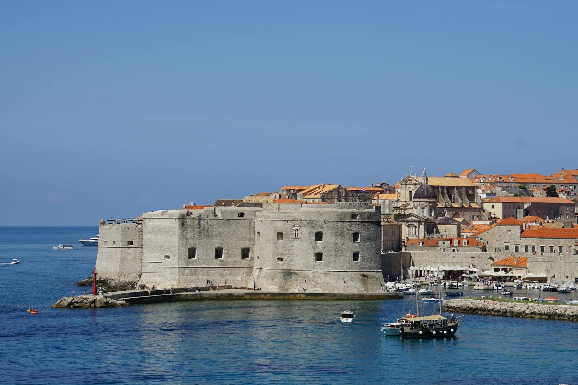 Vista de la Fortaleza de San Juan de Dubrovnik al borde del puerto, rodeada de las cristalinas aguas del Adriático y los tejados naranjas de la ciudad antigua bajo un cielo despejado.