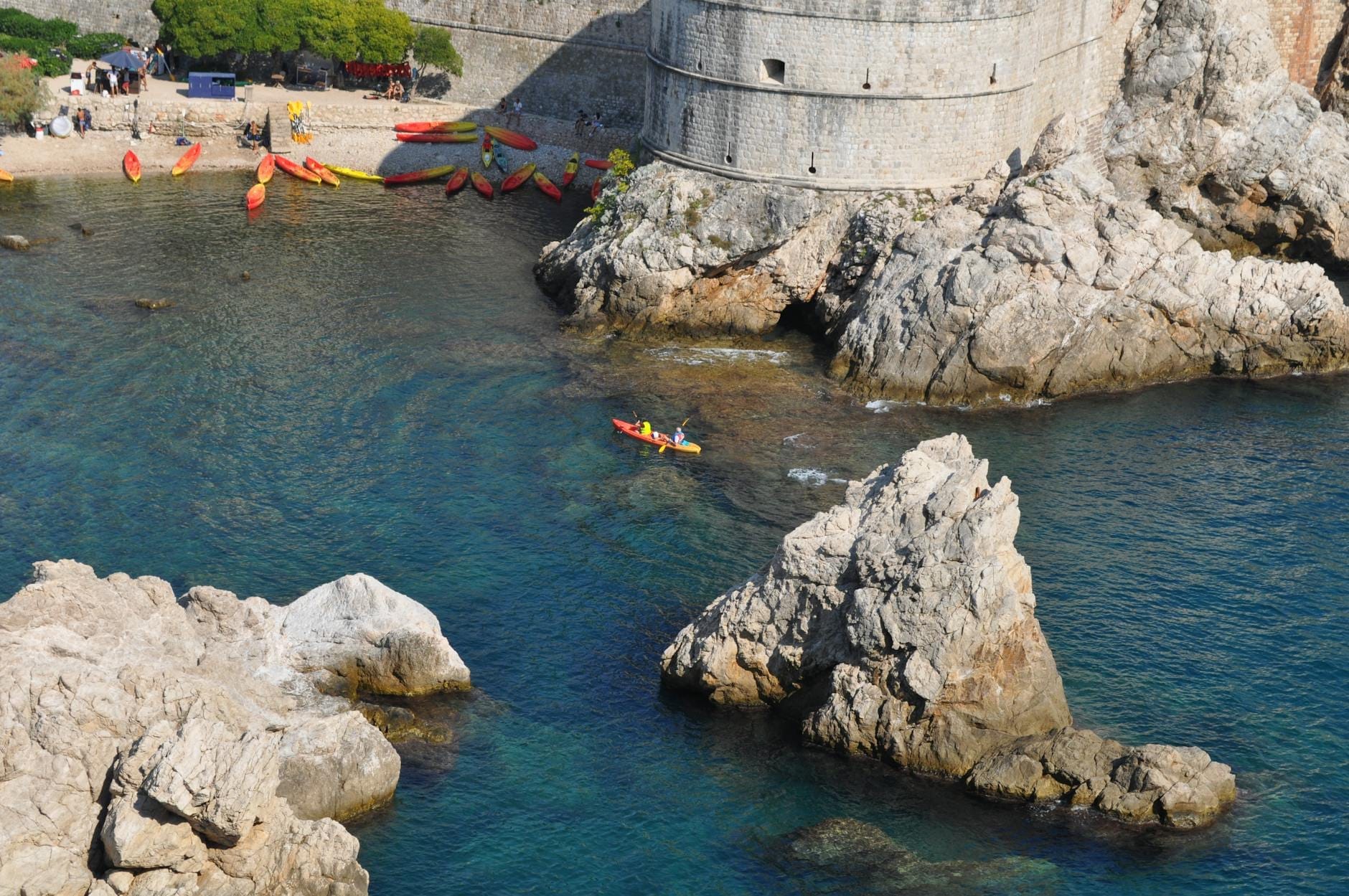 Plage de galets avec kayaks et nageurs au pied des remparts de Dubrovnik, rivage rocheux et eau bleue cristalline visibles.
