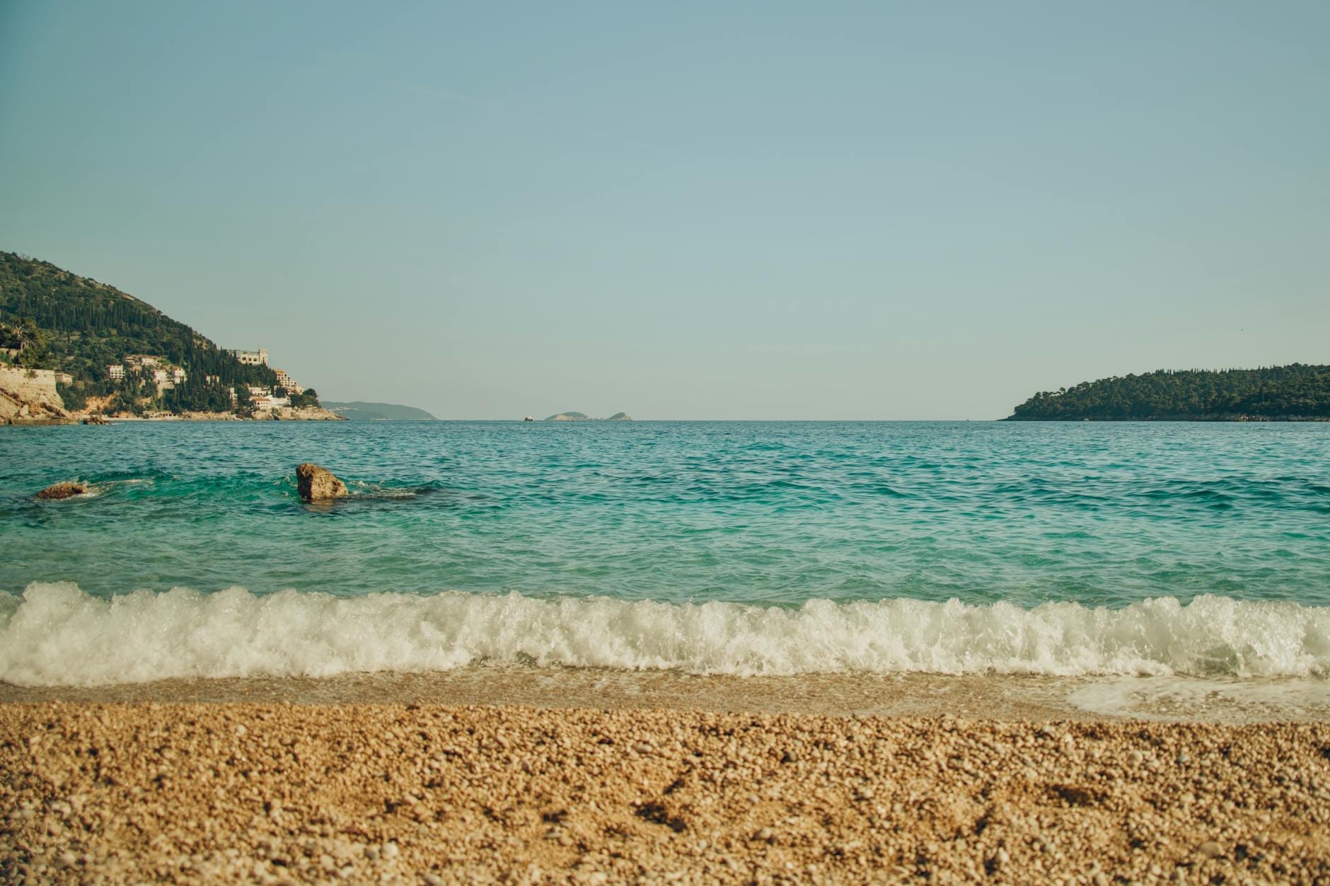 Plage de galets avec une eau turquoise, des vagues douces et une vue sur des collines verdoyantes et des îles près de Dubrovnik par temps clair.
