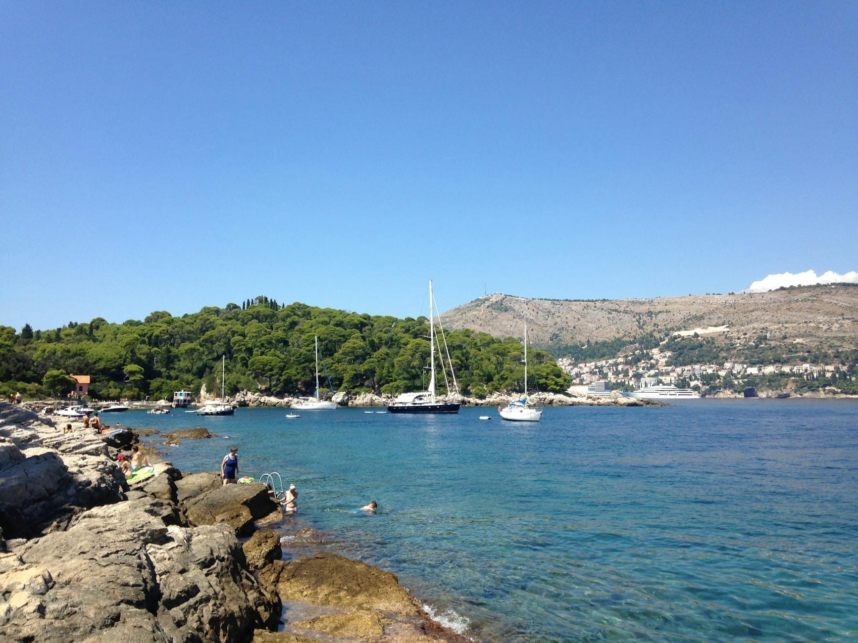 Côte rocheuse avec des personnes se prélassant au soleil et nageant, voiliers ancrés près de l'île de Lokrum avec le paysage urbain de Dubrovnik en arrière-plan.