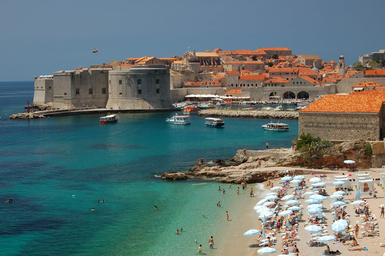 Una playa de arena con bañistas y sombrillas blancas con vistas al agua turquesa y las murallas de la ciudad vieja de Dubrovnik, con barcos cerca de la orilla bajo un cielo despejado.