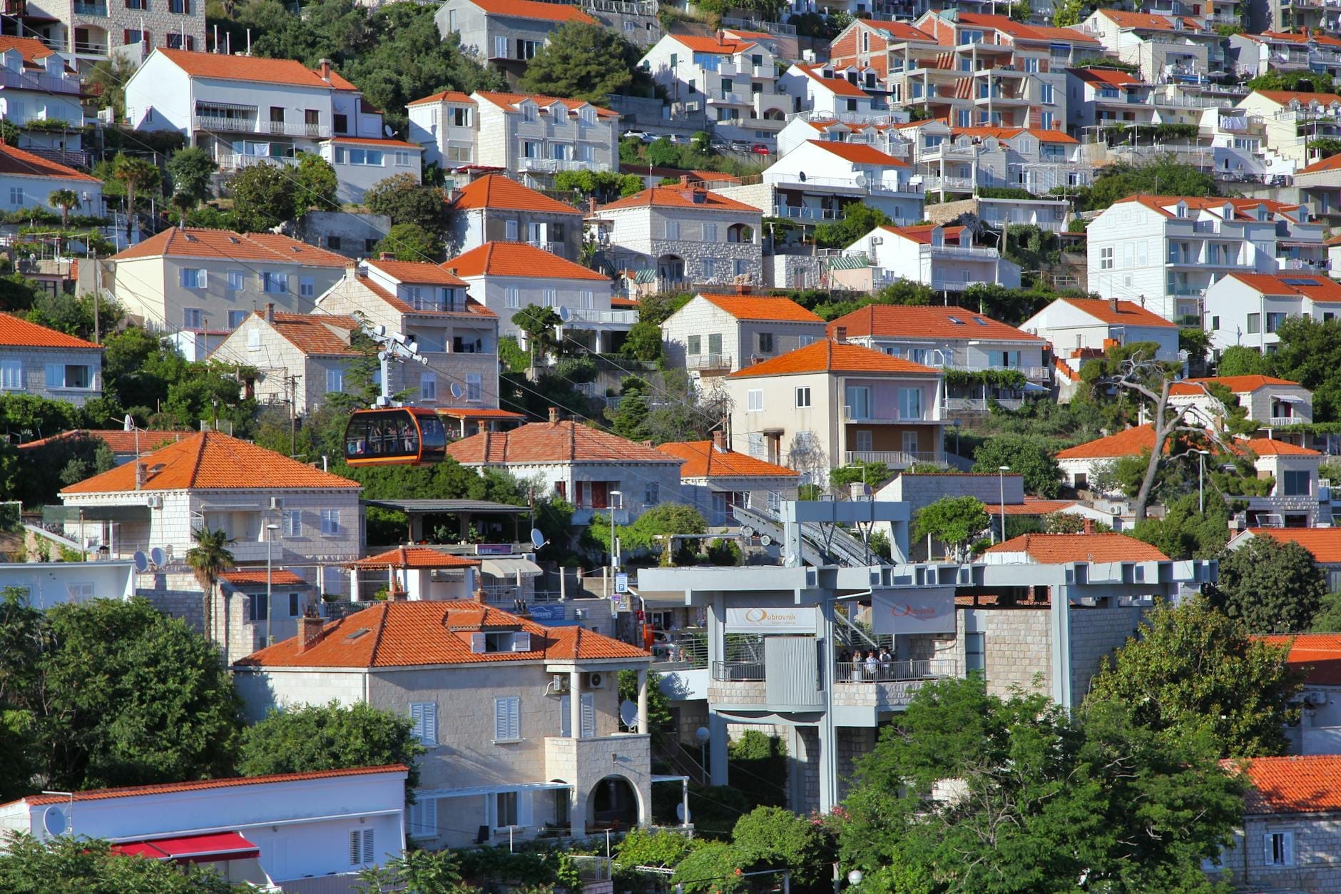 Conjunto de casas de Dubrovnik con tejados naranjas y teleférico pasando por zona residencial en dirección a la estación inferior