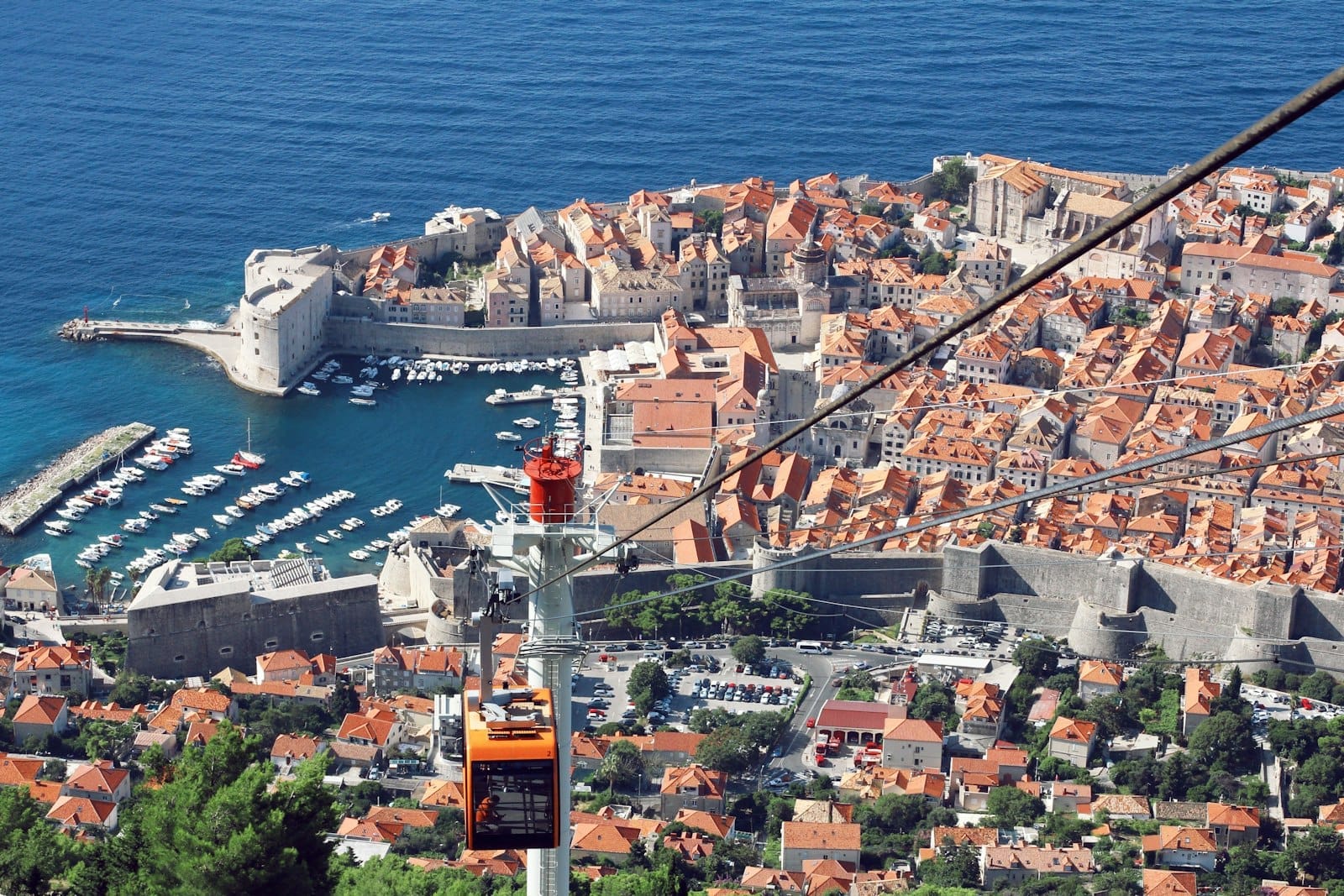 El teleférico naranja de Dubrovnik ascendiendo sobre la Ciudad Vieja, con tejados de terracota, las murallas y el mar Adriático azul al fondo en un día soleado.