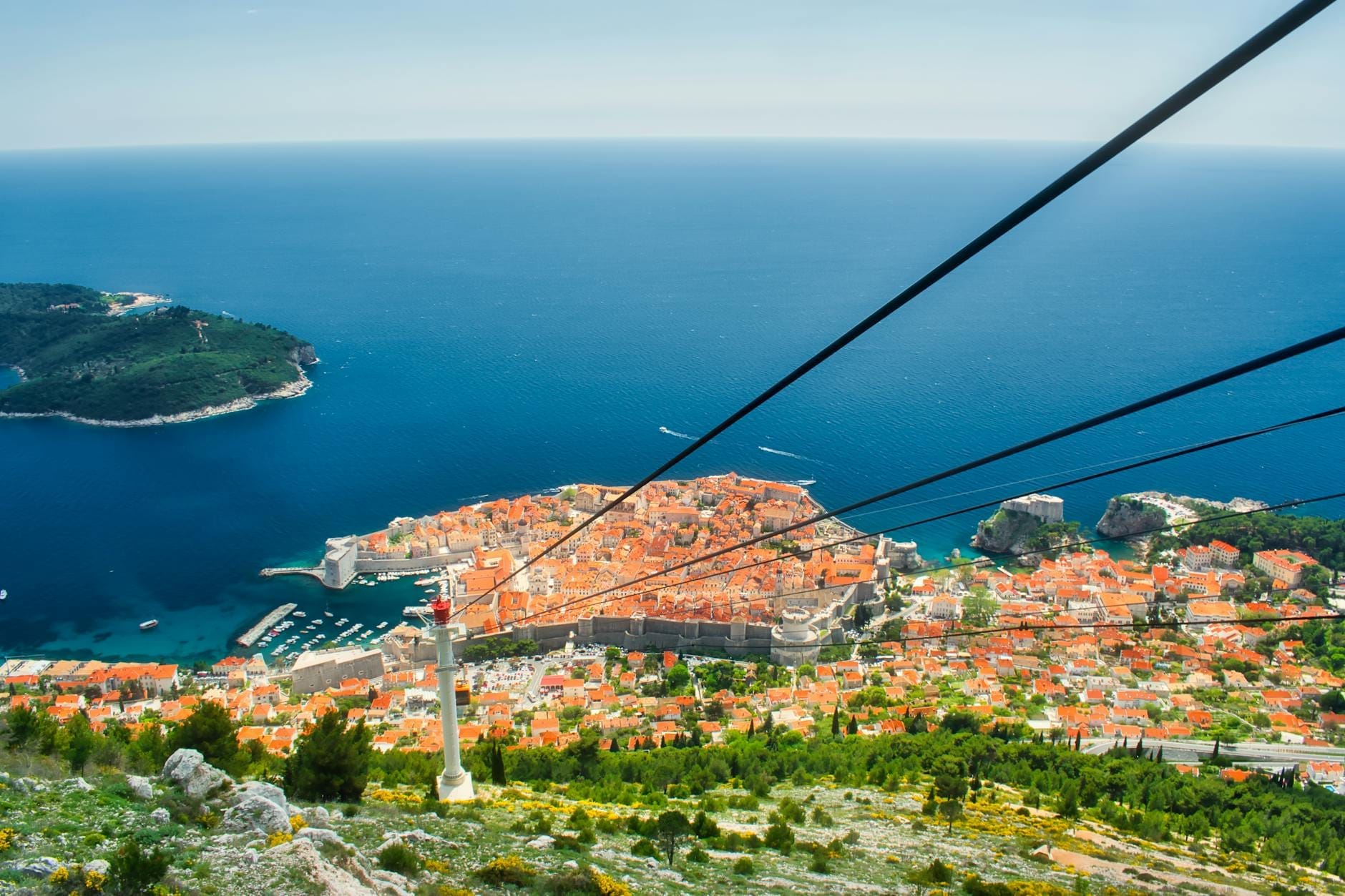 Vista desde lo alto del teleférico de Dubrovnik con la Ciudad Vieja, el mar Adriático, una isla cercana y los tejados de terracota bajo un cielo azul despejado.