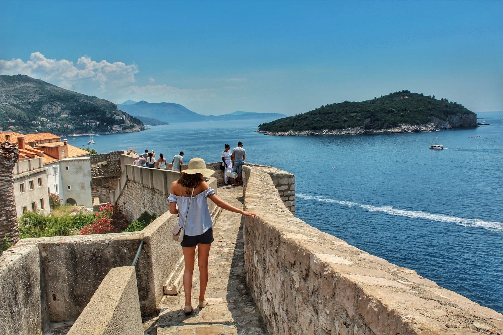 Du khách đi bộ trên Tường thành Dubrovnik với tầm nhìn ra Biển Adriatic và hòn đảo gần đó, dưới thời tiết nắng đẹp rực rỡ.