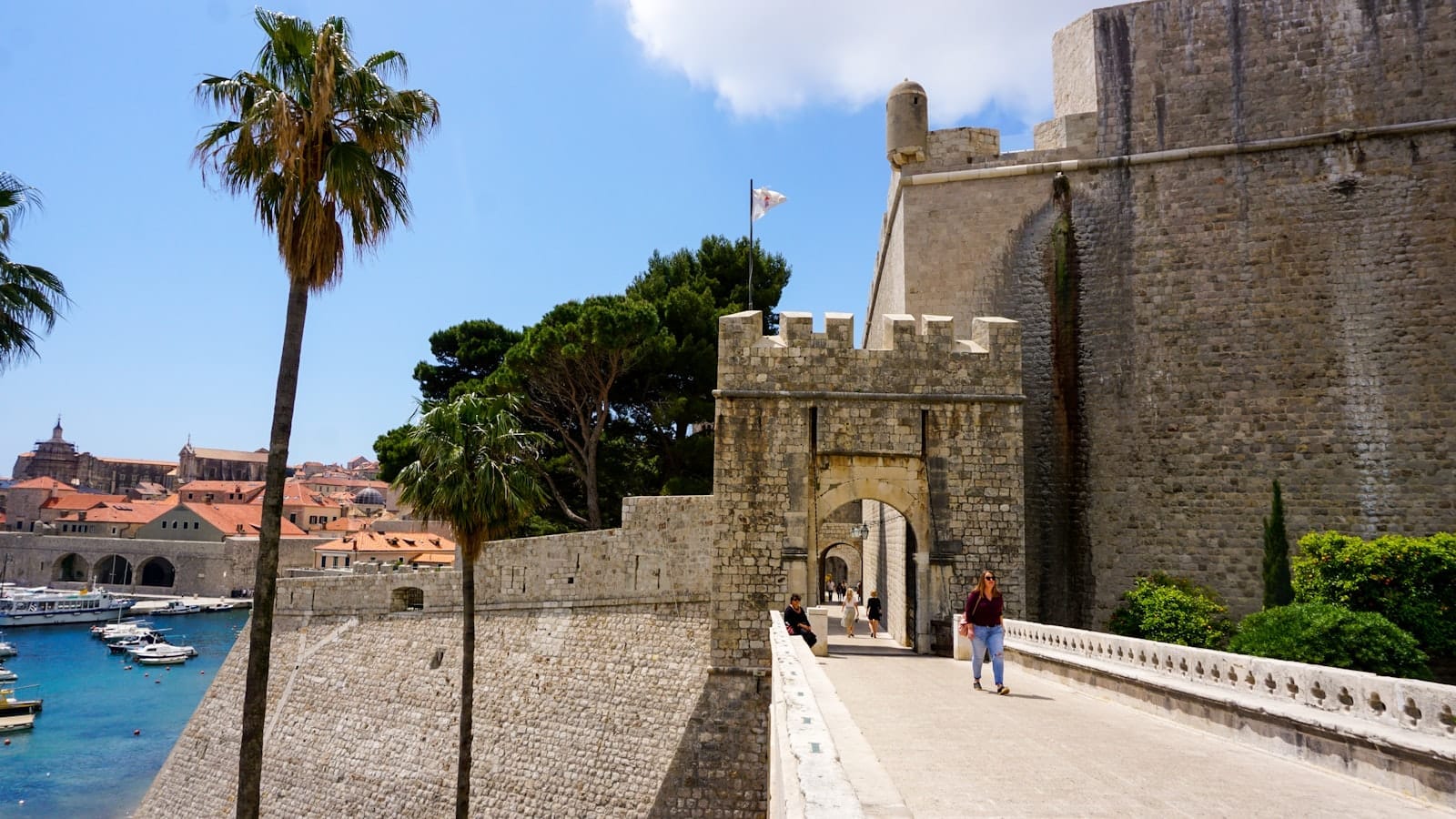 Vista de una puerta de entrada principal a las murallas de la ciudad de Dubrovnik con personas caminando, almenas de piedra y el puerto al fondo.