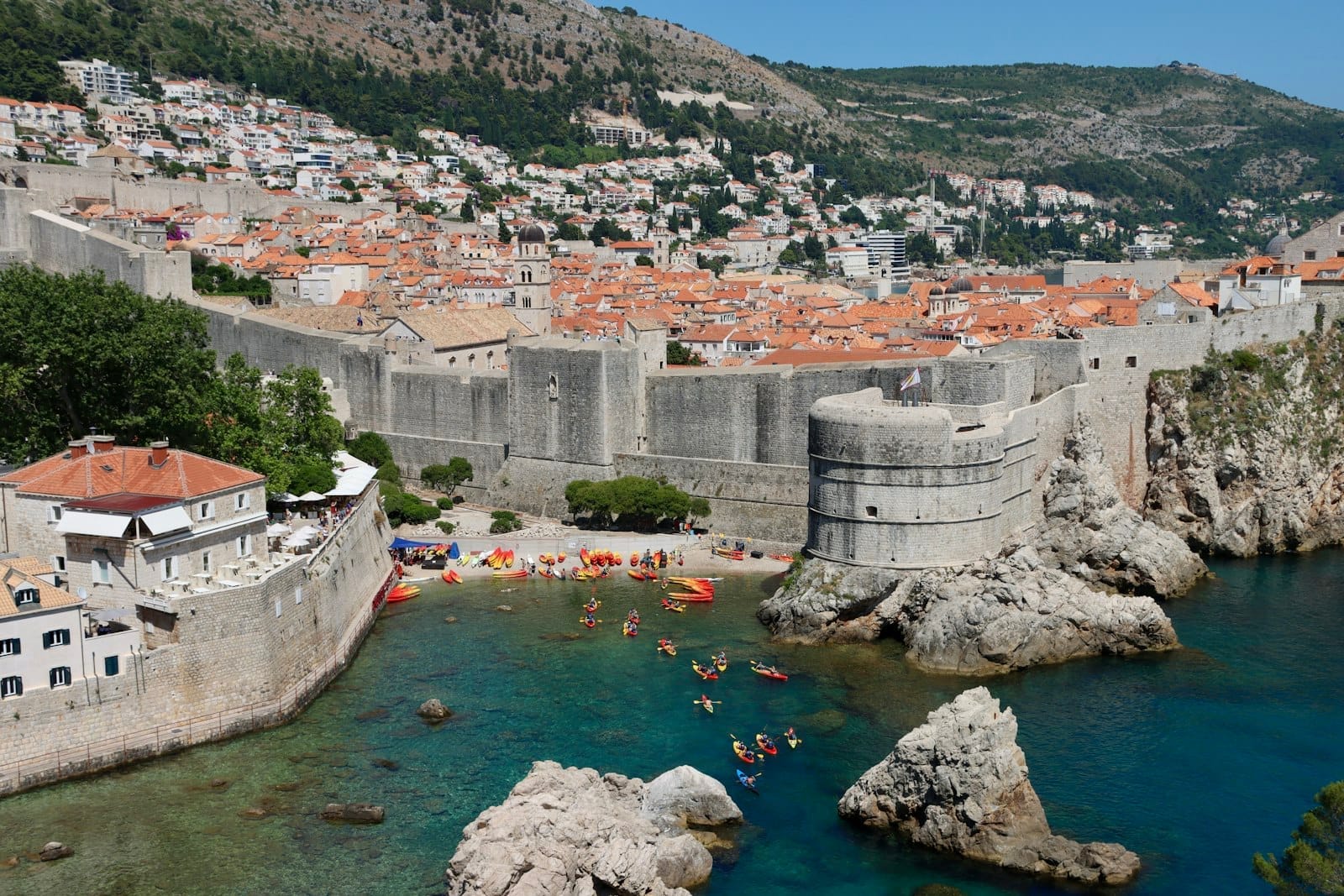 Vista amplia de las murallas de Dubrovnik elevándose sobre el mar, con la ciudad vieja y kayaks a lo largo de la orilla en un día soleado.