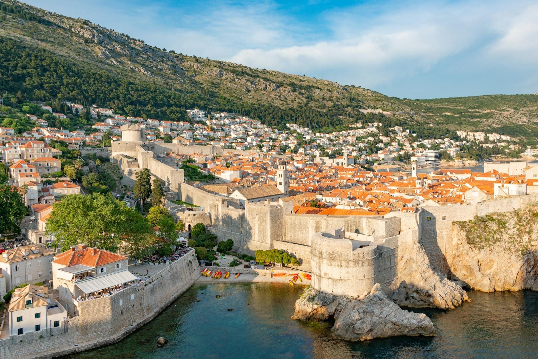 Vista panorámica de las murallas de Dubrovnik rodeando el casco antiguo, con tejados de terracota, mar azul cristalino y colinas verdes en un día soleado.