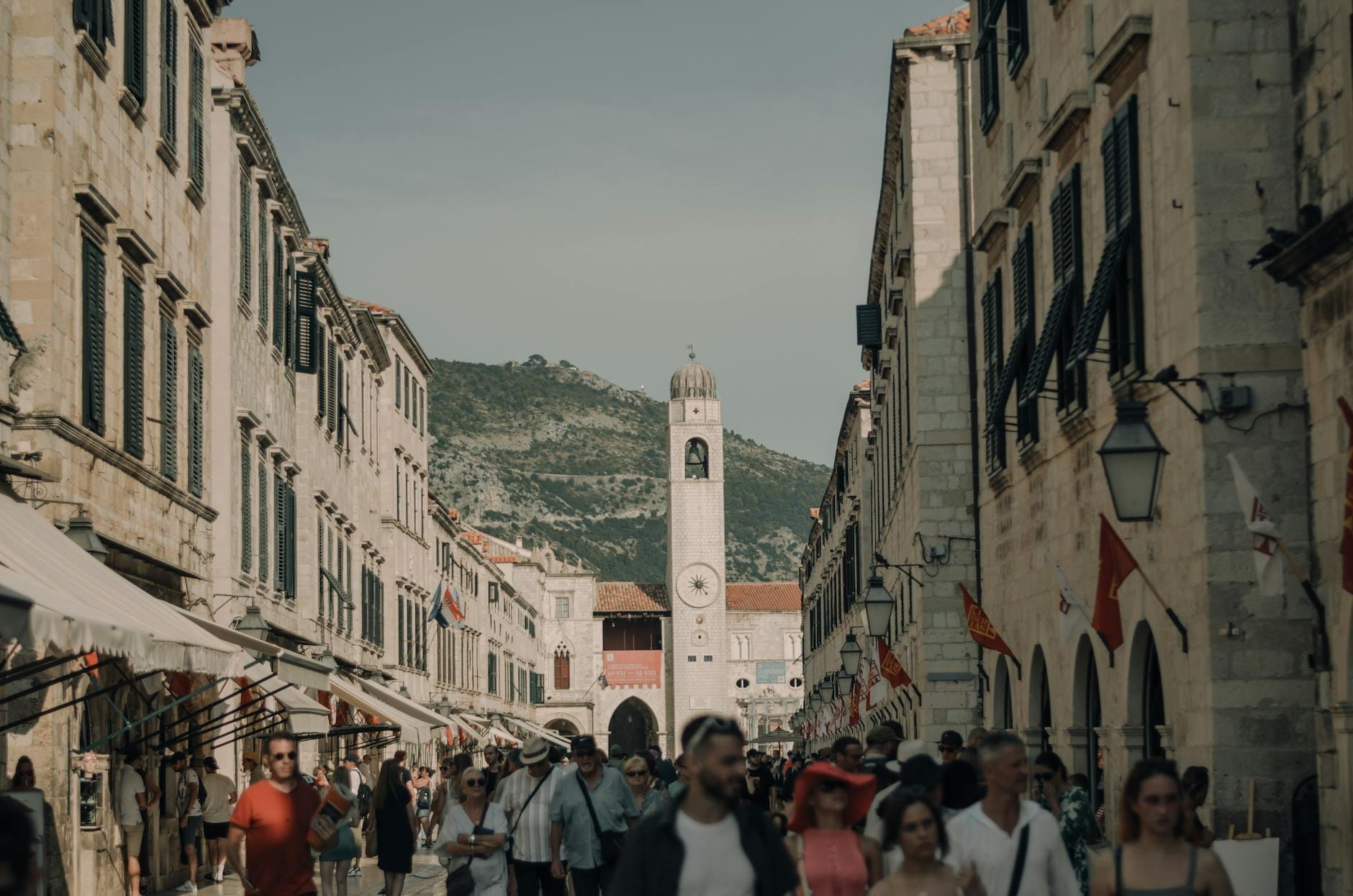 Calle concurrida en el casco antiguo de Dubrovnik con multitudes, históricos edificios de piedra caliza y una torre del reloj al fondo.