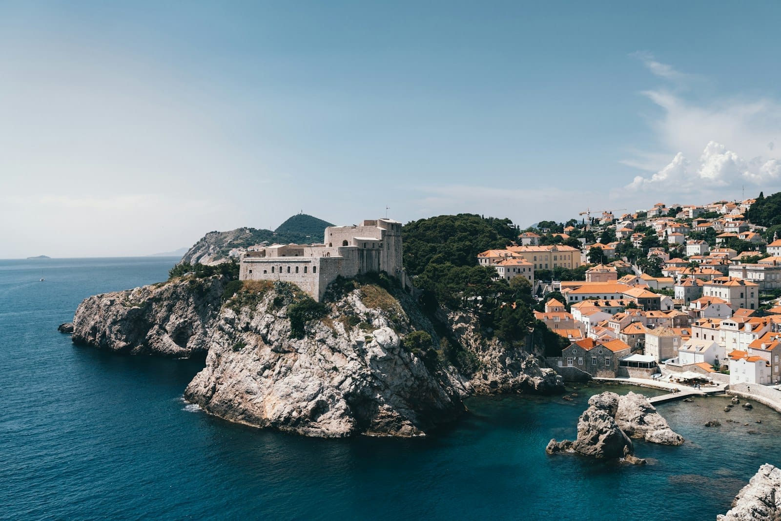 Vista de las murallas de piedra y el casco antiguo de Dubrovnik en la costa adriática, con cielos despejados, acantilados y tejados de terracota.