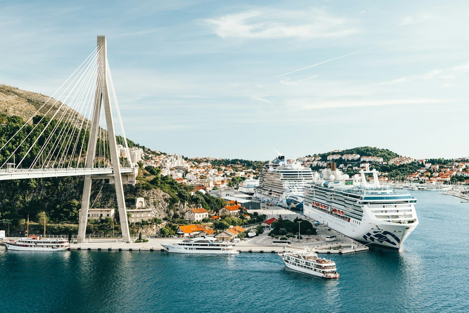 Grandes cruceros atracados en el puerto principal de Dubrovnik cerca de un puente atirantado, con edificios de la ciudad y colinas al fondo.
