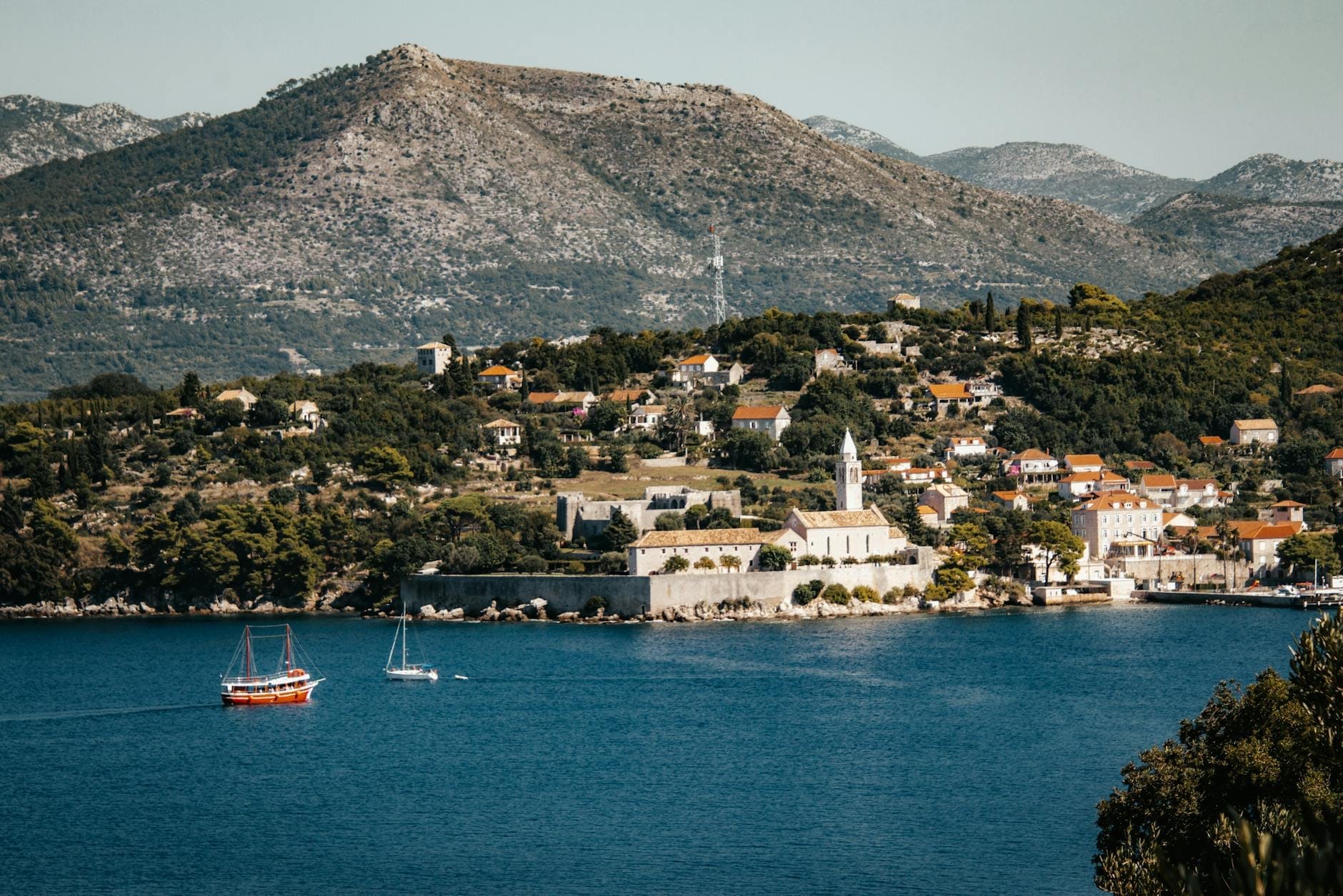 Vue pittoresque d'un village côtier près de Dubrovnik avec des bateaux sur une eau bleue, des bâtiments blancs, des montagnes en arrière-plan et une végétation luxuriante.