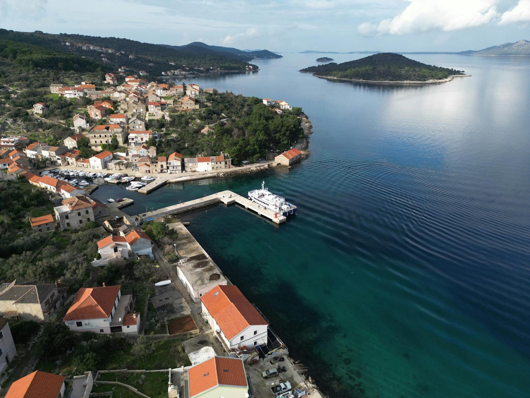 Vue aérienne d'une petite ville portuaire avec des maisons aux toits rouges et des collines verdoyantes, un ferry à la jetée et les îles Elaphiti dans une mer bleue calme.
