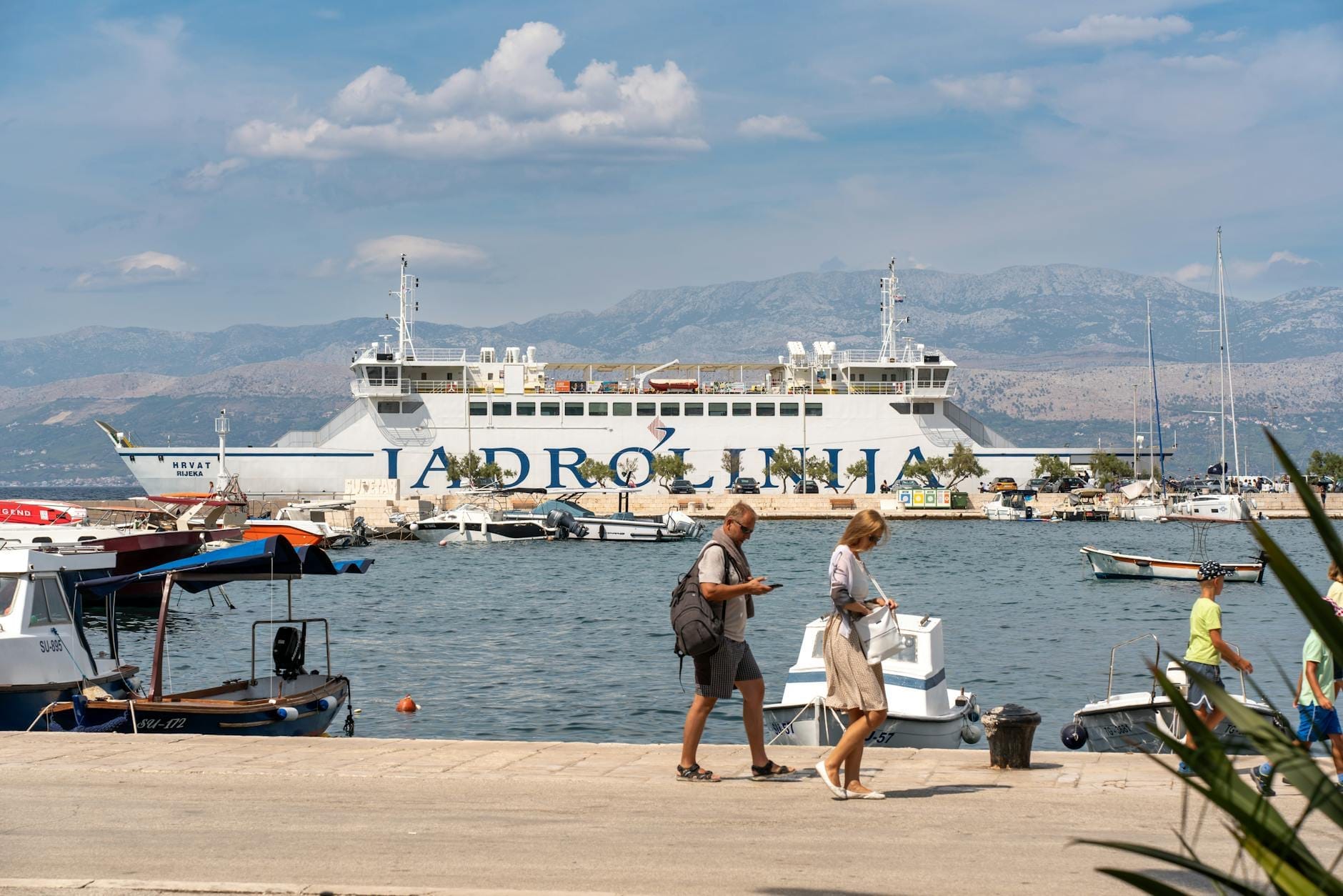 Grand ferry Jadrolinija amarré dans un port avec des personnes se promenant sur la jetée et des bateaux en arrière-plan.