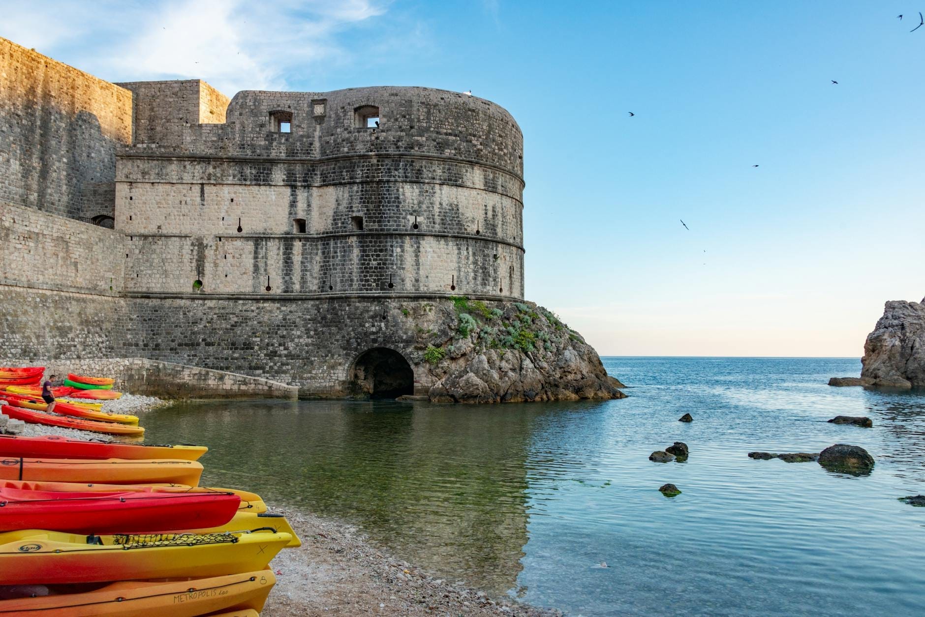 Kayaks de colores vivos alineados en la orilla junto a las antiguas murallas de piedra de Dubrovnik, con el mar Adriático y el cielo azul de fondo.