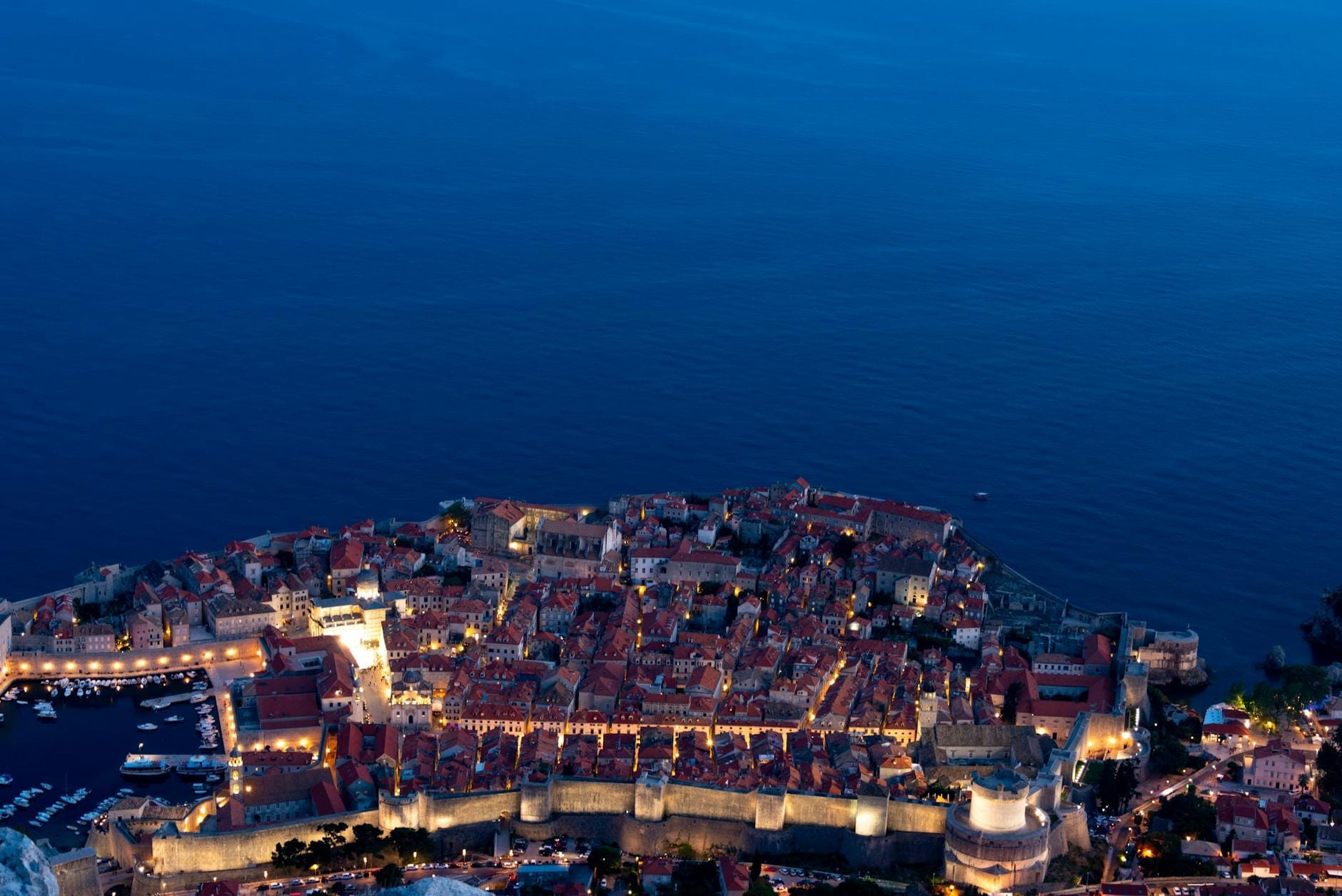 Vista aérea al atardecer de la ciudad vieja de Dubrovnik con las murallas medievales iluminadas y el tranquilo mar Adriático al fondo, evocando el ambiente nocturno de la ciudad.