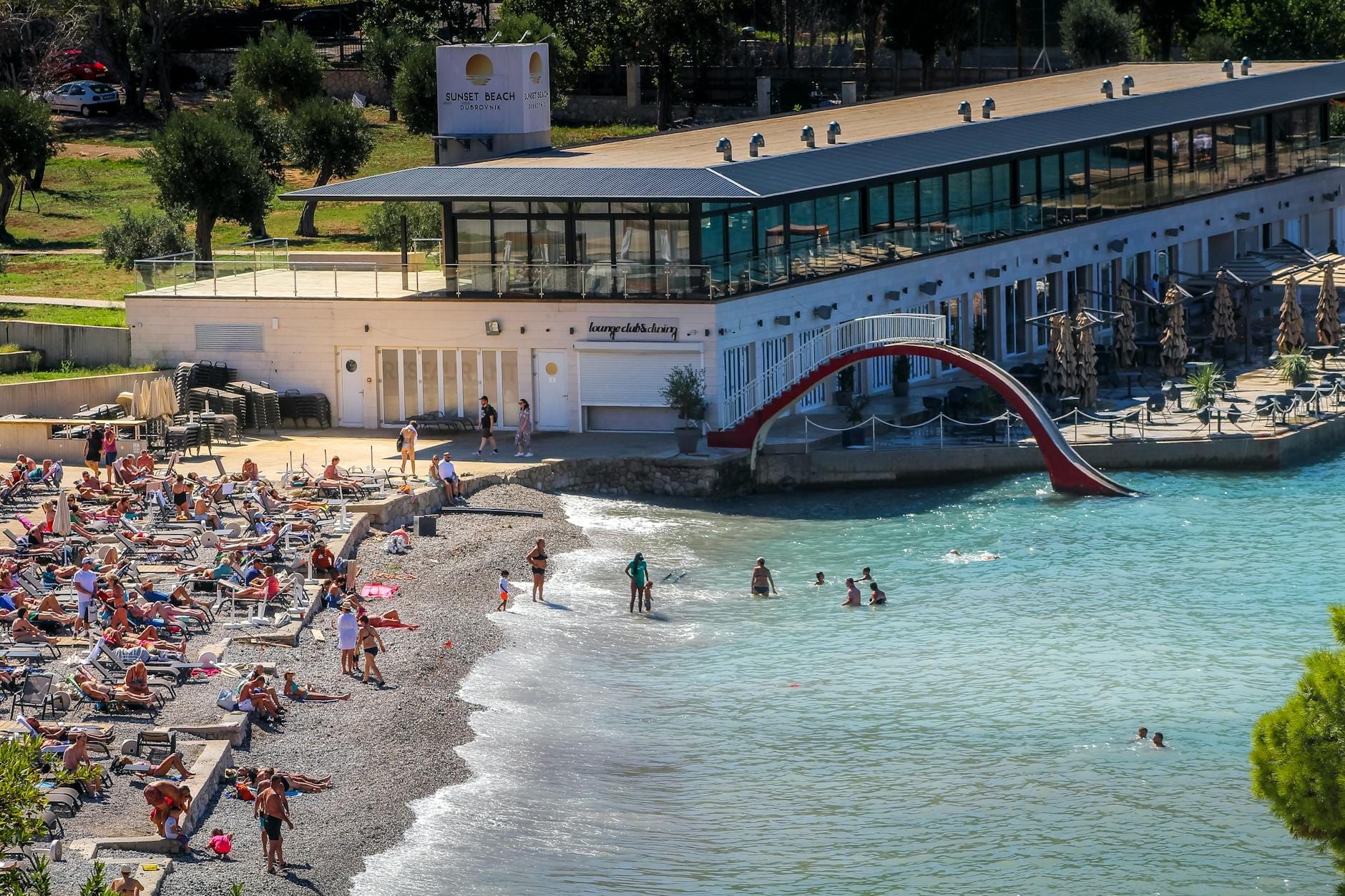Beach club animé de Dubrovnik avec des transats, des personnes se relaxant, nageant et un bar moderne avec vue sur la mer cristalline.