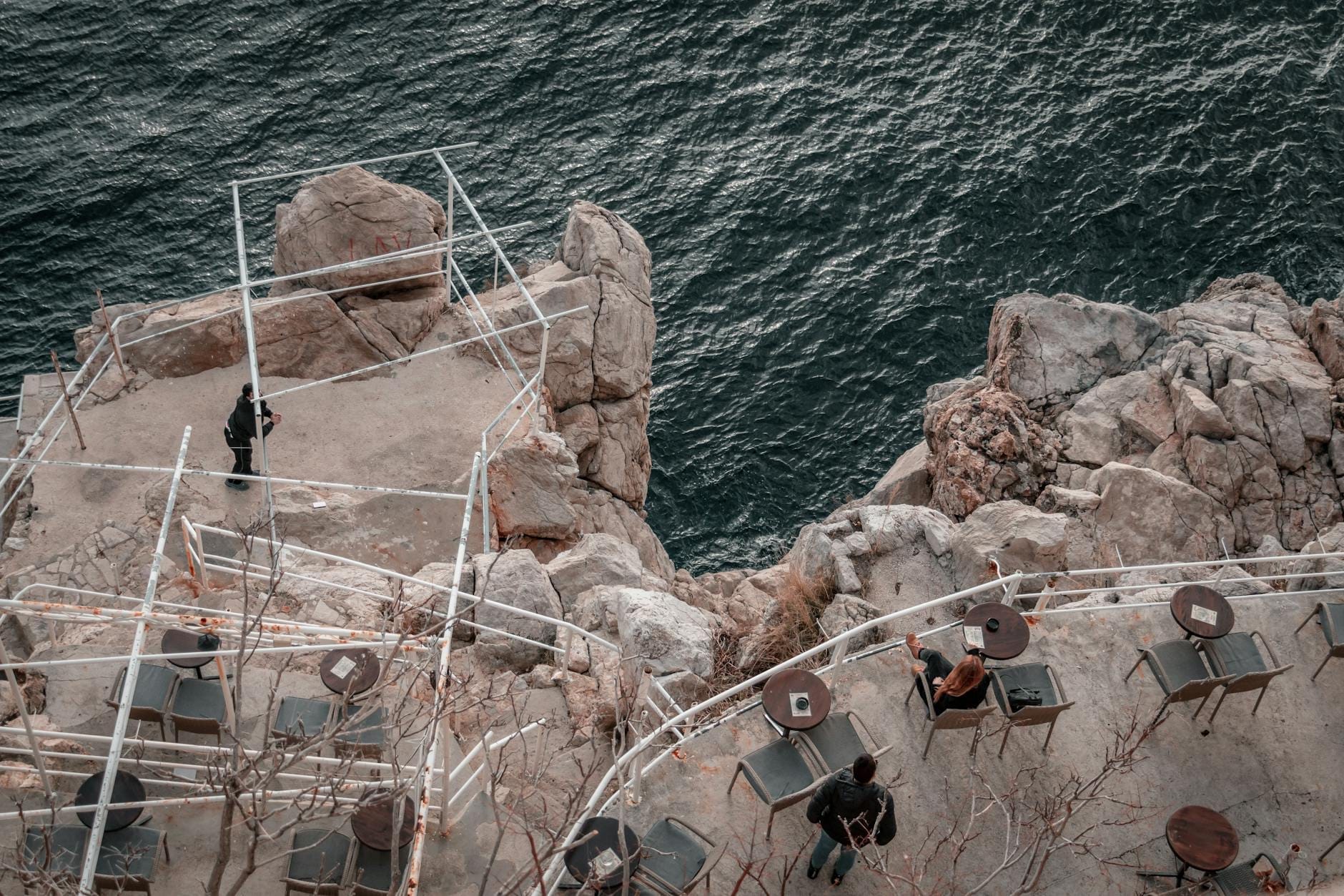 Tables et chaises sur la terrasse d'un bar à flanc de falaise surplombant l'océan, avec des personnes profitant des vues spectaculaires sur les corniches rocheuses à Dubrovnik.