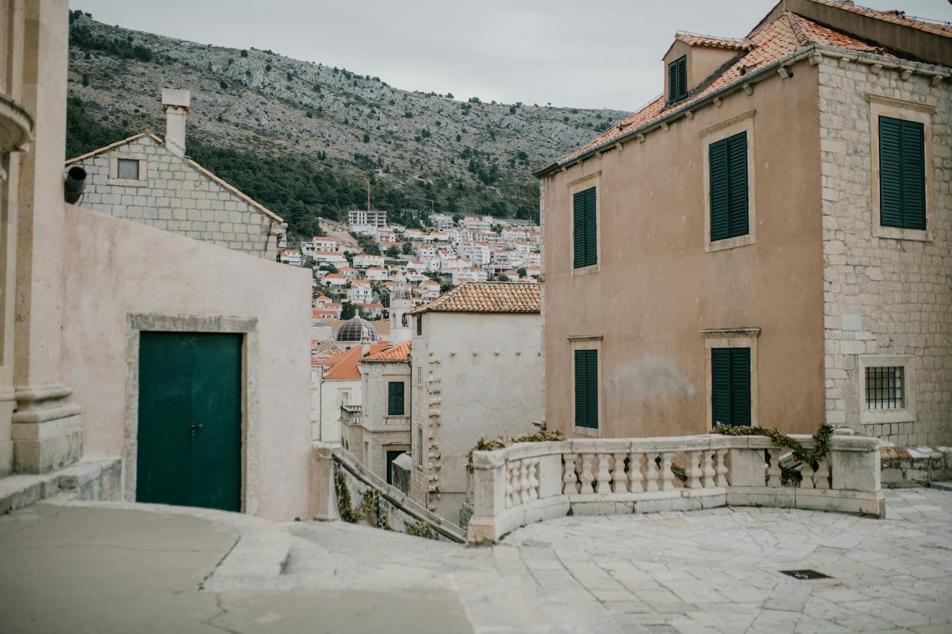 Rue résidentielle calme à Dubrovnik avec des maisons en pierre, des volets verts et des habitations sur la colline en arrière-plan, suggérant une atmosphère de quartier local.